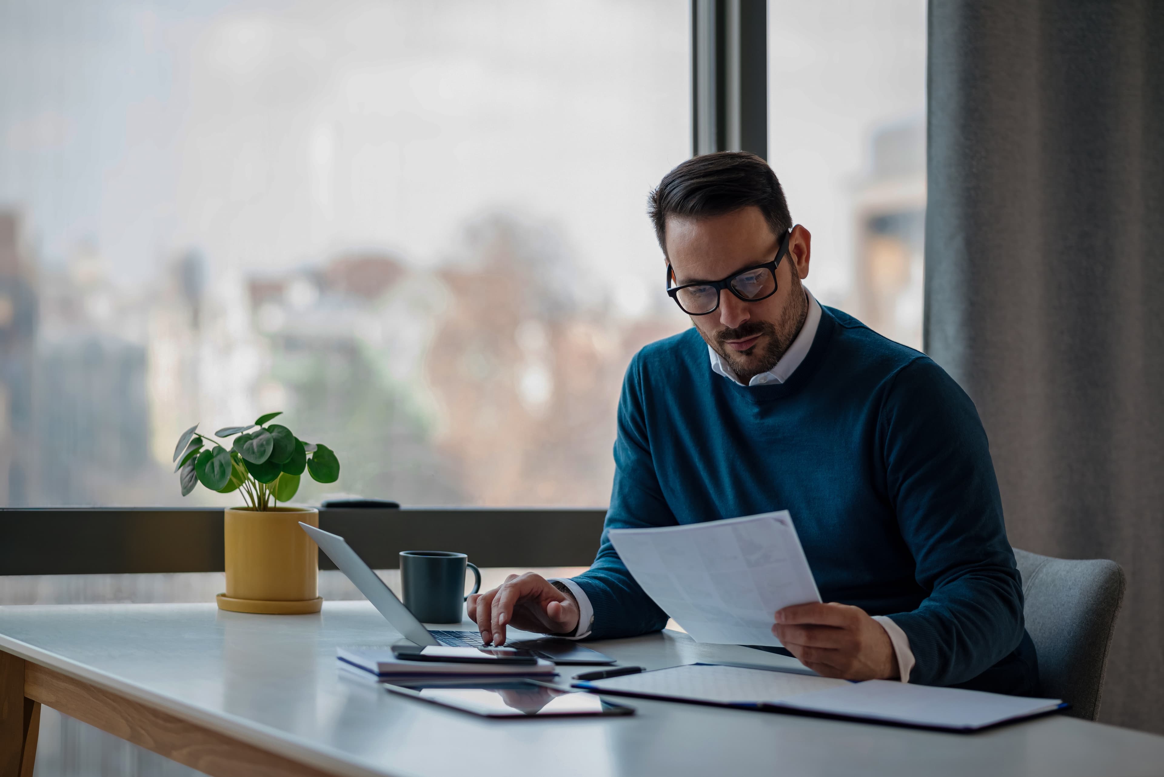Mann mit Laptop und Unterlagen an Schreibtisch. Im Hintergrund Fenster mit Blick über die Stadt.