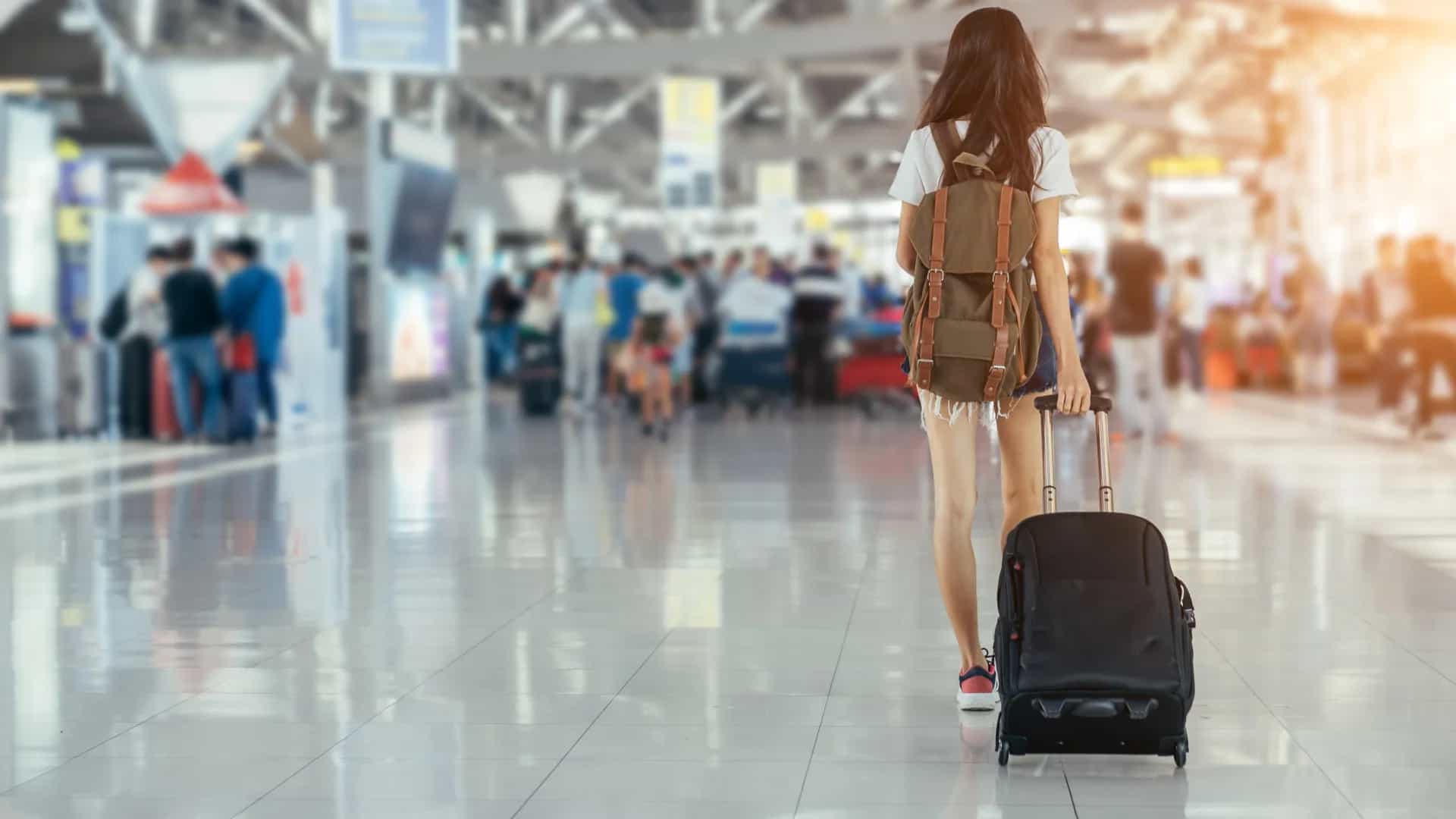 Frau mit Koffer an Flughafen. Im Hintergrund Flughafenbetrieb.